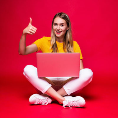 Happy young curly beautiful woman sitting on the floor with crossed legs and using laptop on red background.
